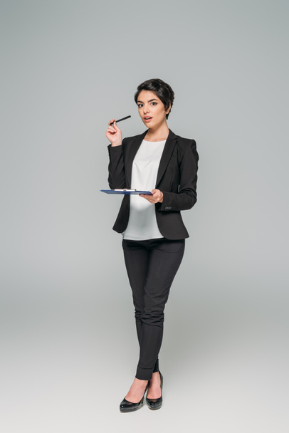 pretty mixed race businesswoman holding clipboard and looking at camera on grey background - Foto, imagen