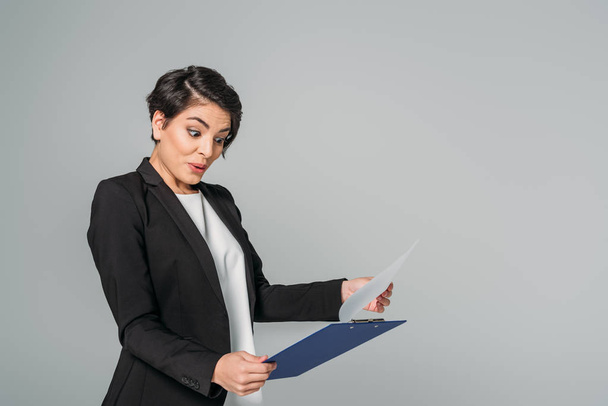 surprised mixed race businesswoman looking at clipboard and gesturing isolated on grey - Foto, imagen