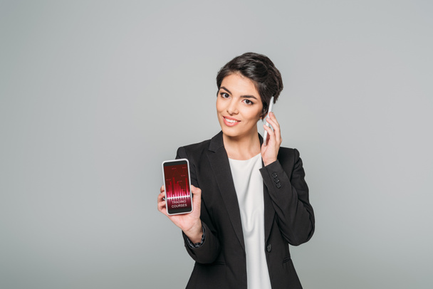 beautiful mixed race businesswoman showing smartphone with trading courses app on screen while talking on smartphone isolated on grey - Photo, Image
