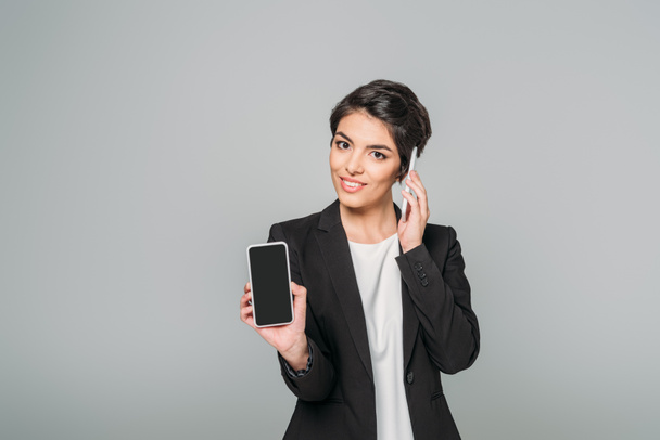 young mixed race businesswoman talking on smartphone while holding smartphone with blank screen isolated on grey. - Zdjęcie, obraz