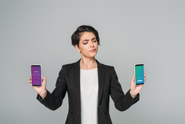 KYIV, UKRAINE - APRIL 24, 2019: Skeptical mixed race businesswoman holding smartphones with Instagram and Twitter apps on screen isolated on grey. - 写真・画像