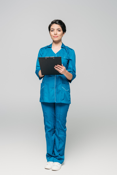 young mixed race doctor holding clipboard while looking at camera on grey background - Foto, imagen