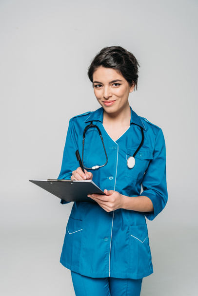 cheerful mixed race doctor with stethoscope writing on clipboard while looking at camera isolated on grey - Foto, Imagem