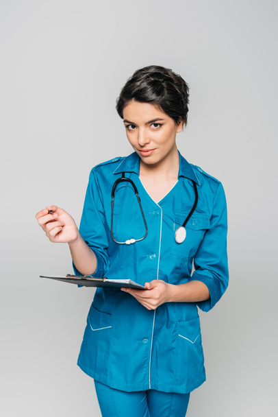 attractive mixed race doctor writing on clipboard and looking at camera isolated on grey - Photo, Image