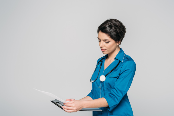 attentive mixed race doctor with stethoscope looking at clipboard isolated on grey - Photo, image