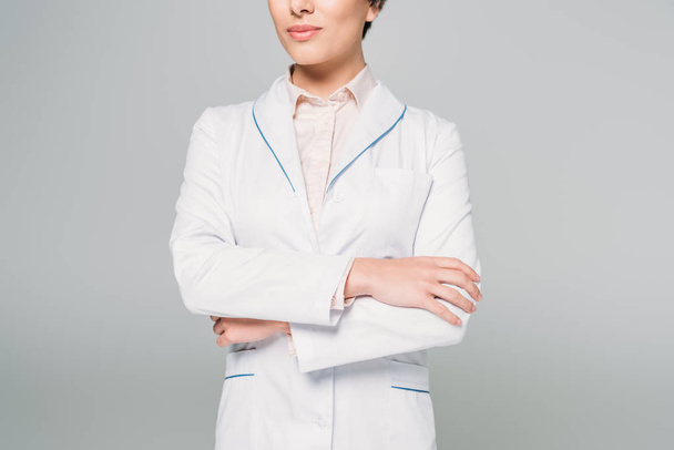 cropped shot of mixed race doctor in white coat posing at camera with crossed arms isolated on grey - Foto, imagen