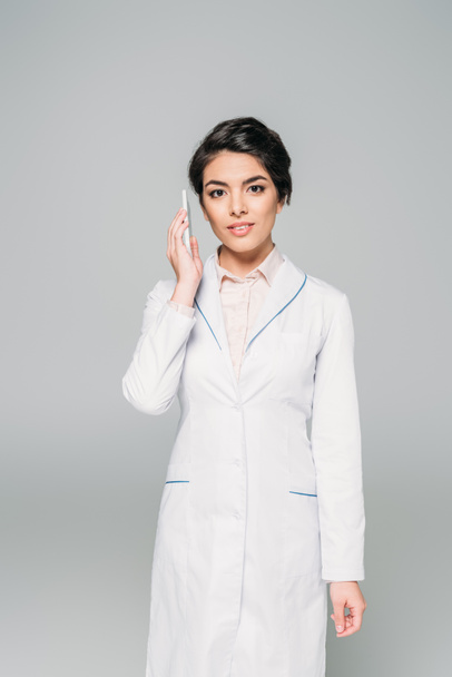 pretty mixed race doctor talking on smartphone and looking at camera isolated on grey - Photo, Image
