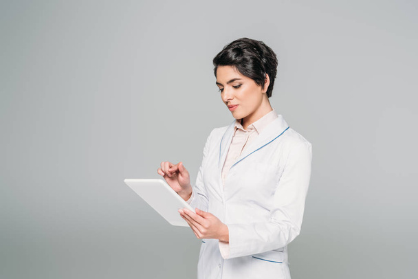 attraente medico di razza mista in camice bianco utilizzando tablet digitale isolato su grigio
 - Foto, immagini