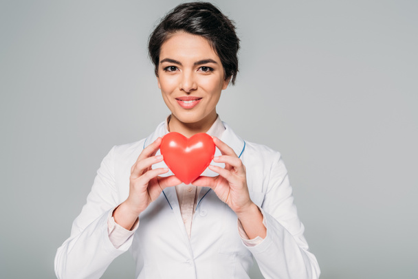 Veselý smíšený závodní lékař držící srdeční model při úsměvu na kameře na šedé - Fotografie, Obrázek