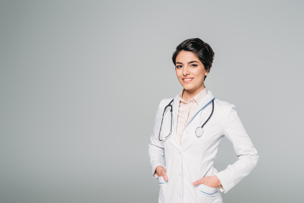 cheerful mixed race doctor with stethoscope posing at camera with hands in pockets isolated on grey - Fotografie, Obrázek