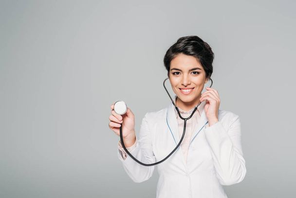 Veselý smíšený závodní lékař v bílém plášti za použití stetoskop izolovaného na šedé - Fotografie, Obrázek