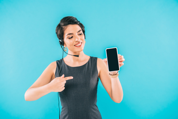 sonriente operador de centro de llamadas de raza mixta apuntando con el dedo en el teléfono inteligente con pantalla en blanco aislado en azul
 - Foto, imagen