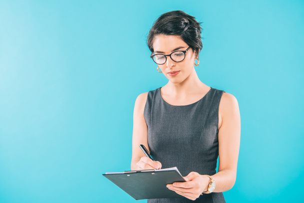 attentive mixed race businesswoman in glasses writing in clipboard isolated on blue - Photo, Image