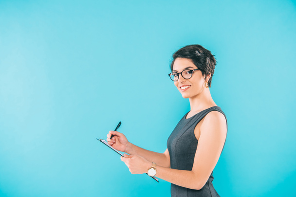 attractive mixed race  businesswoman writing on clipboard while looking at camera isolated on blue - Foto, Bild