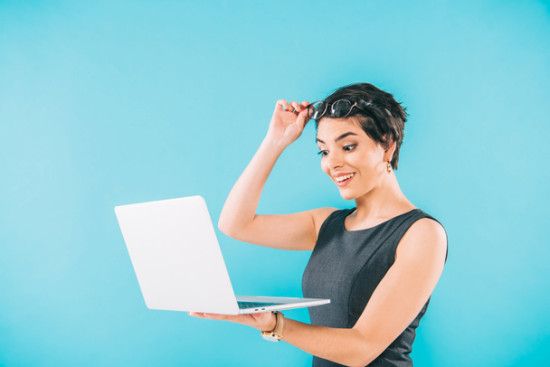 surprised mixed race businesswoman touching glasses while using laptop isolated on blue - Foto, Imagem