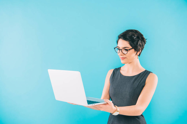 beautiful mixed race businesswoman in glasses using laptop isolated on blue - Фото, зображення