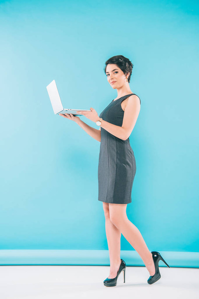 fashionable mixed race businesswoman holding laptop and looking at camera on blue background - Фото, зображення