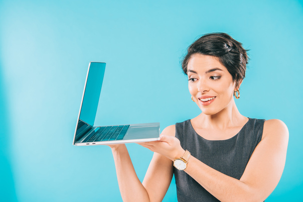 vrolijke gemengde race zakenvrouw in grijze jurk houden laptop op blauwe achtergrond - Foto, afbeelding