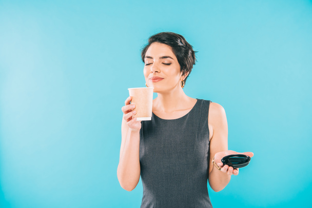 attraktive Frau mit gemischter Rasse genießt den Geschmack von Kaffee mit geschlossenen Augen, während sie eine Papptasse auf blauem Hintergrund hält - Foto, Bild