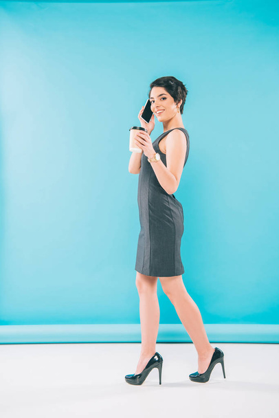 mujer de raza mixta de moda posando en la cámara mientras habla en el teléfono inteligente y sosteniendo la taza de papel sobre fondo azul
 - Foto, imagen