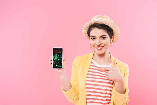smiling mixed race woman pointing with finger at smartphone with marketing analysis app on screen isolated on pink - Photo, Image