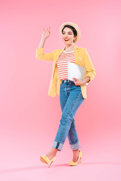 excited mixed race woman walking with laptop and waving hand on pink background - Foto, imagen
