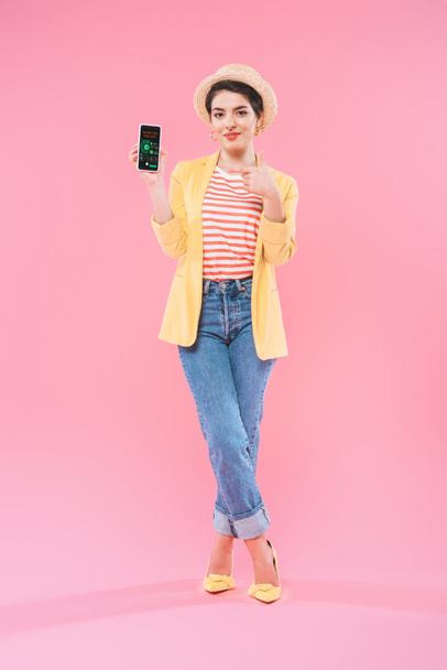 mujer de raza bastante mixta en ropa brillante que muestra el teléfono inteligente con la aplicación de análisis de marketing en pantalla sobre fondo rosa
 - Foto, Imagen