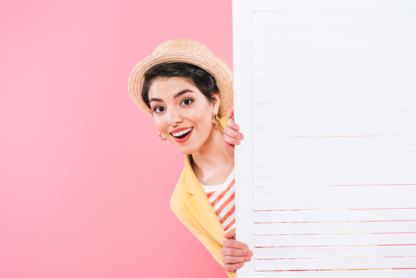 cheerful mixed race woman looking out folding screen on pink background - Foto, Imagen