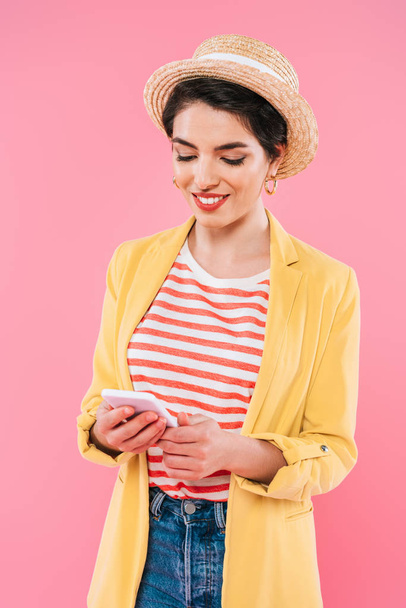 hymyilevä sekarotuinen nainen kirkkaissa vaatteissa ja olkihattu käyttäen älypuhelin eristetty vaaleanpunainen
 - Valokuva, kuva