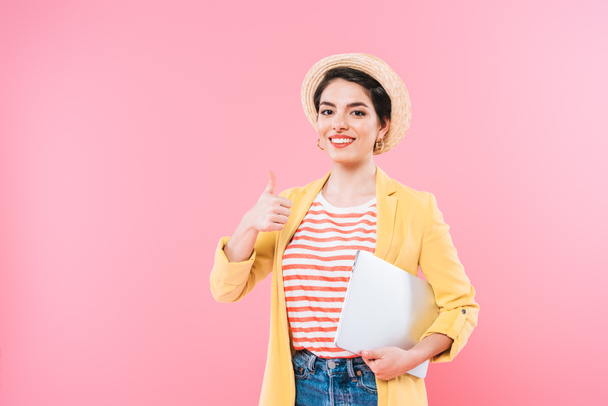 cheerful mixed race woman holding laptop and showing thumb up isolated on pink - Фото, изображение