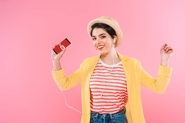 KYIV, UKRAINE - APRIL 24, 2019: Cheerful mixed race woman holding smartphone with Youtube app on screen and listening music in earphone isolated on pink. - Zdjęcie, obraz