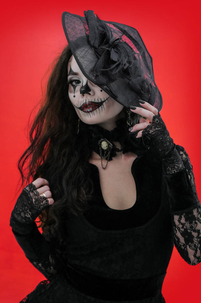 Portret van een jonge brunette met make-up in de stijl van Halloween in zwarte kleren en hoed. Verschrikkelijk meisje met enge mond en ogen, extreme Body-Art. Het beeld van een heks of een boze clown. - Foto, afbeelding