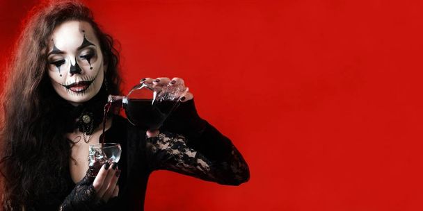 A Halloween-style brunette wearing black clothes pours a red liquid that looks like blood into a skull-shaped glass dish. A blank for invitation card, banner or poster on the theme of a terrible party - Foto, immagini