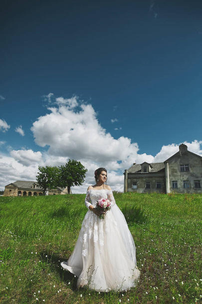 Beautiful model woman in stylish wedding dress holding a bouquet of flowers and posing on the green grass with an ancient building at the background - Fotografie, Obrázek