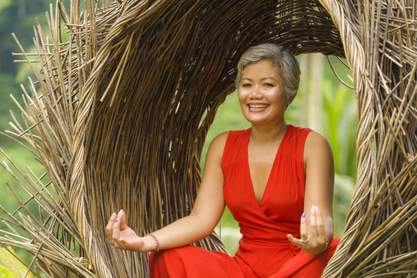 attractive and happy 40s or 50s middle aged Asian woman in classy and beautiful red dress practicing yoga relaxation and meditation in tropical jungle - Fotoğraf, Görsel