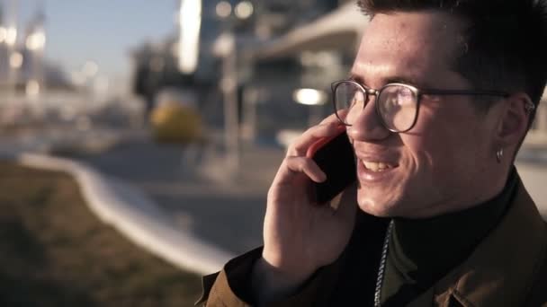 Cheerful, smiling man in stylish glasses and earing is talking using his smarphone while sitting outside on parapet near the seaside. Close up. Slow motion - Felvétel, videó