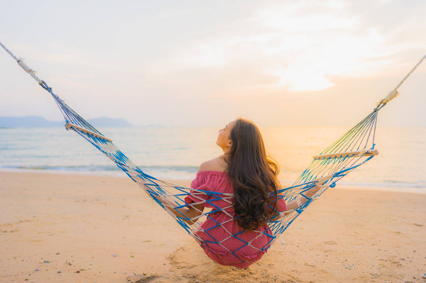 Portrait beautiful young asian woman sitting on the hammock with smile happy neary beach sea and ocean for leisure travel and vacation at sunset time - Photo, Image