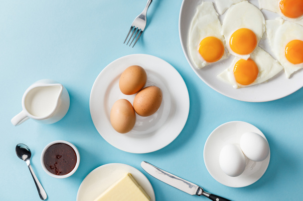top view of boiled and fried eggs, butter, jam on white plates, milk, fork, spoon and knife on blue background - Photo, Image