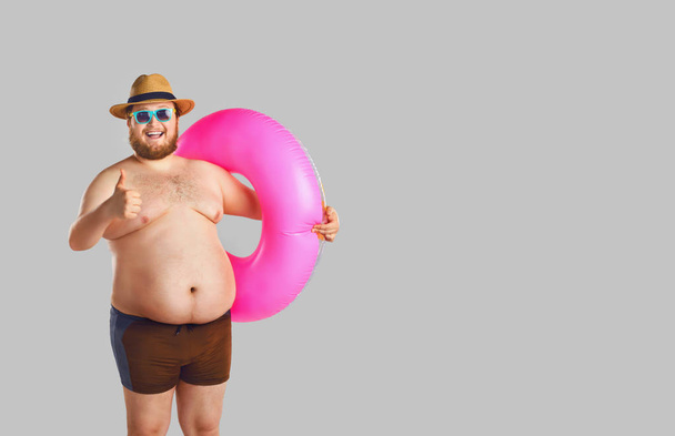 Fat funny man in swimming trunks with an inflatable ring on a gray background. - Photo, Image