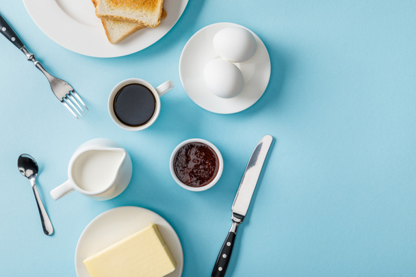 top view of fresh eggs, jam, coffee, cutlery, butter and two toasts on white plates on blue background - Photo, Image