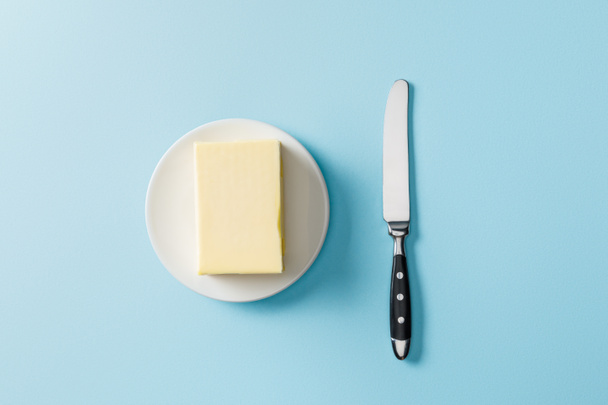 top view of butter on white plate and knife on blue background - Zdjęcie, obraz
