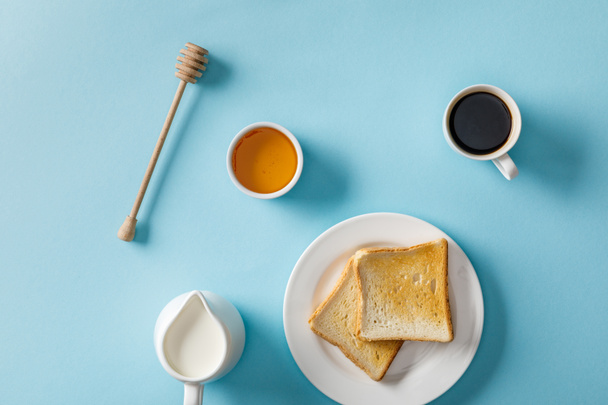 top view of honey in bowl, wooden dipper, coffee, milk and two toasts on white plate on blue background - 写真・画像