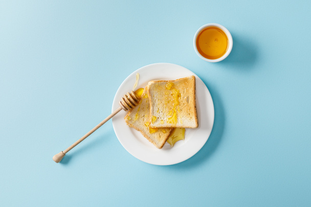 top view of toasts with honey and wooden dipper on white plate on blue background  - Fotoğraf, Görsel