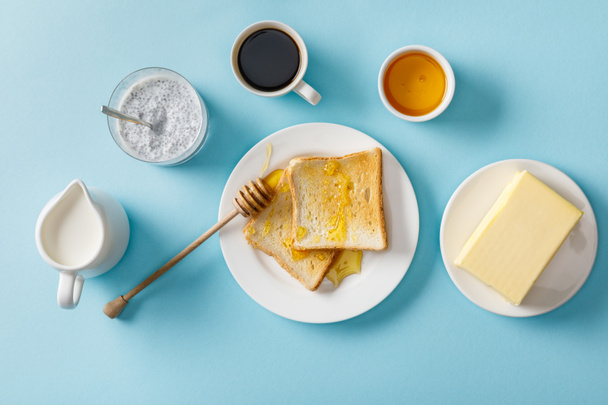 top view of coffee, milk, yogurt with chia seeds, butter, honey, toasts with honey and wooden dipper on white plates on blue background - Foto, Imagen