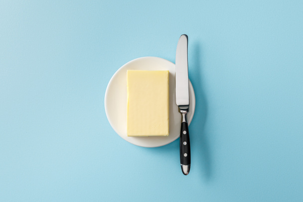 Top zicht op boter en mes op witte plaat op blauwe achtergrond - Foto, afbeelding