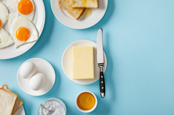 vista superior de mantequilla, tostadas, huevos frescos y fritos en platos blancos, yogur con semillas de chía y tazón con miel sobre fondo azul
 - Foto, Imagen
