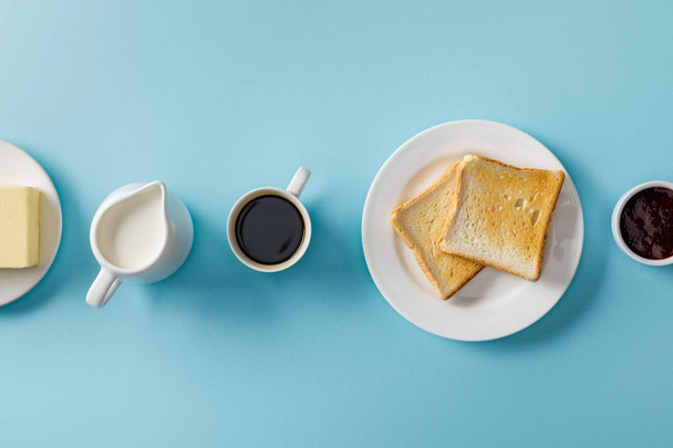 Top View van kopje koffie, melk, boter, jam en twee toast op witte plaat op blauwe achtergrond - Foto, afbeelding