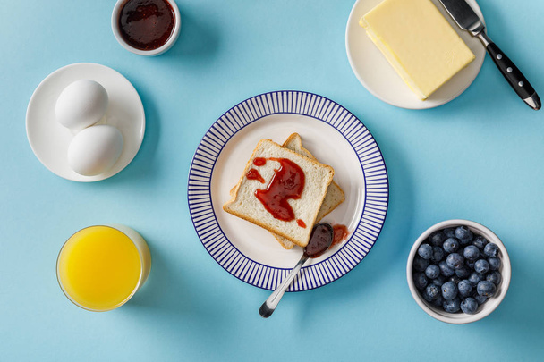 top view of toasts with jam, orange juice, boiled eggs, butter and blueberries on blue background - Zdjęcie, obraz
