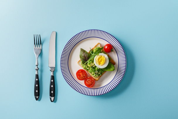 top view of toast with guacamole, boiled egg cherry tomatoes, fork and knife on blue background - Fotó, kép