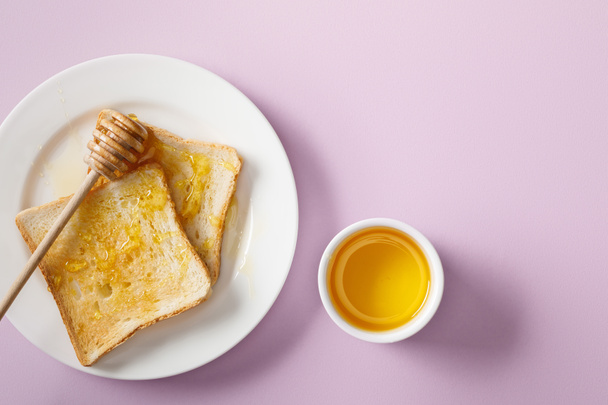 top view of bowl, toasts with honey on white plate and wooden dipper on violet background - Foto, Imagem